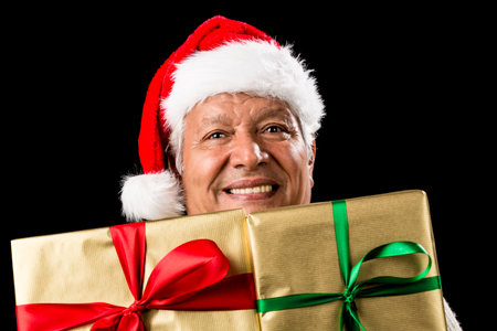 Smiling male senior peeping across two foot-long gifts that cover his chest and chin. Both presents wrapped golden with green and red bows. Father Christmas hat. Gift giving theme isolated on black.の写真素材
