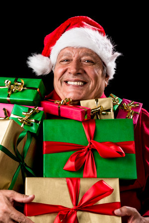 Jovial male senior with broad grin is peering across a load of presents that he is carrying against his chest. Gifts wrapped in plain red, green, magenta and gold. Red Father Christmas cap and coat.の写真素材