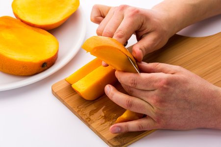 Closeup of a man?s hands peeling one of four mango wedges over a bamboo cutting board. The blade of a short kitchen knife is separating the yellow, juicy fruit pulp from its skin. White background.の写真素材