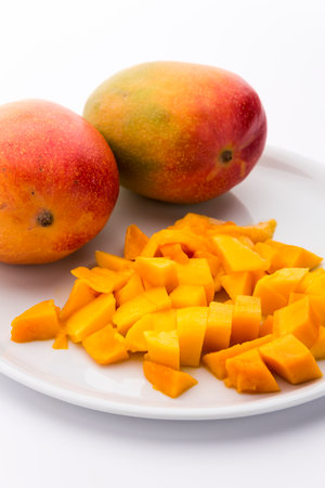Orange yellow cubes of juicy mango fruit pulp presented on a white plate. Two entire mangos in the back. Vertical orientation. Copy space. White background. Medium wide depth of field.の写真素材