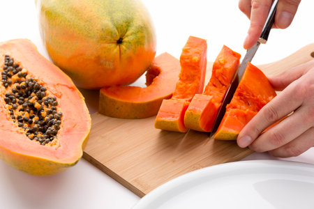 Halved papaya fruit being cut into slices with a sharp kitchen knife on a wooden cutting board. Half a pawpaw fruit and an entire papaya placed next to it. Orange colored fruit pulp. White background.の写真素材