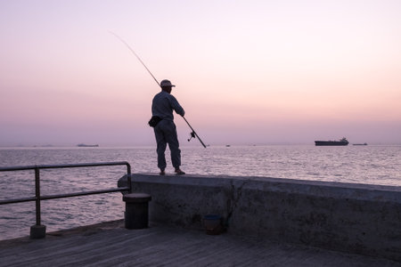 Elderly man silhouette standing on stone pier and throwing fishing rod into the sea on seascape background at beautiful sunset. Boats and remote islands in haze on horizon line.の写真素材