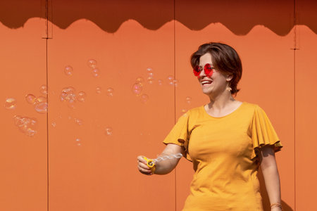 Beautiful young brunette woman in sunglasses having fun blowing soap bubbles. Metal texture orange...