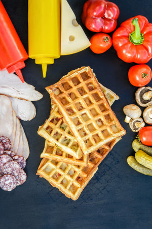 Traditional belgian waffles with cheese and vegetables on a black background. Cucumber. Ham. Mushrooms. Tomato. Salami. Cheese. Pepper. Ketchup. Mustard. Copy space. Flat lay. Top viewの写真素材
