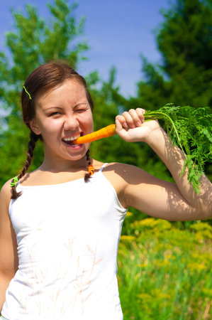 Young smiling woman bites wth effort fresh orange organic carrot in gardenの写真素材