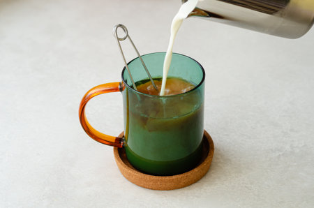 Close up of masala black tea brewed in green glass cup with tea infuser on cork coaster, milk poured from steel jug. Warming spiced drink for autumn and winter.