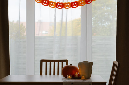 Minimal autumn table decor with pumpkin, white vase and candles by window with paper pumpkin garland for Halloweenの写真素材