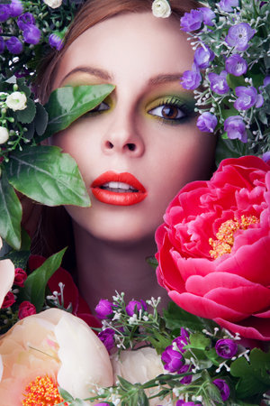 Closeup art portrait of a young cute woman over blue background. Perfect bright makeup. Red lips. Red straight hair. Fashion attractive beautiful girl. With many flowers.の写真素材