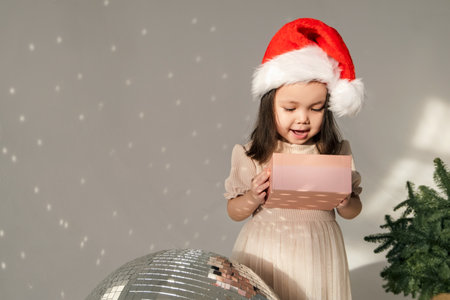 Little cute Asian girl toddler stands near the disco ball and spruce branches. She holds a gift box. High quality horizontal photoの写真素材