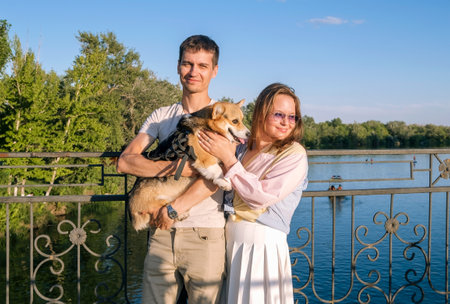 Happy young couple walking in the park with a small corgi dog. Woman and man standing on the bridge in summer. Beautiful family, fun, emotions. Loveの写真素材