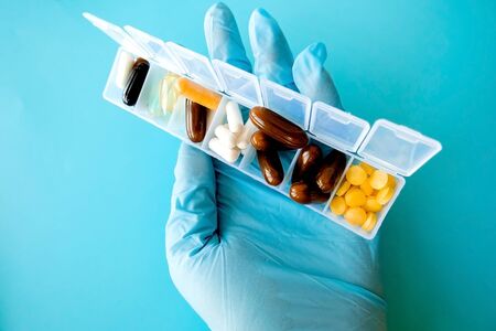 A hand with a medical glove holds multi-colored capsules, a pill box with medicines on a blue...