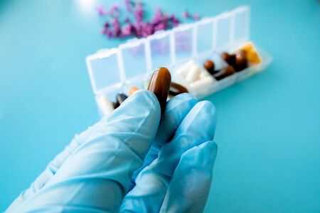 A hand with a medical glove holds multi-colored capsules, a pill box with medicines on a blue backgroundの写真素材