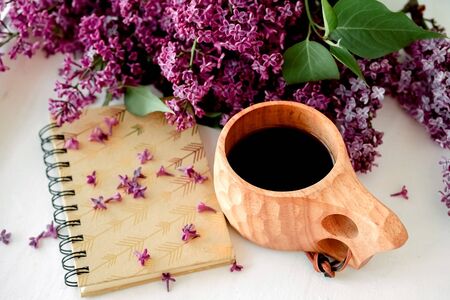 Wooden finnish cup with coffee on the table. Nearby lies a closed notebook for notes and a bouquet of lilacs. Good morning and good mood, planning a day at breakfast, concept
の写真素材