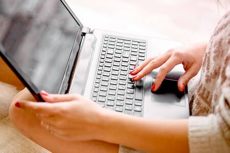 girl in a light knitted sweater sits near the window and holds a laptop. Female hands print on a...