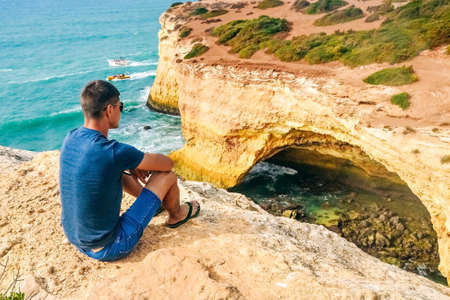 one slender handsome guy with glasses sits on stones on a rock in Portugal. back view of the oceanの写真素材