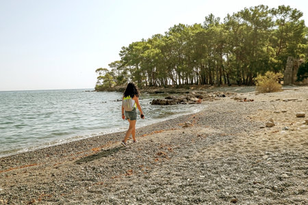 slender brunette girl in denim shorts and a white tank top. woman tourist with a backpack walking along the lagoon beach, view from the back, phaselis old townの写真素材