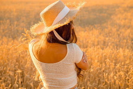 slender beautiful girl in a straw hat in the field, back view. golden ears of wheat, harvest season...
