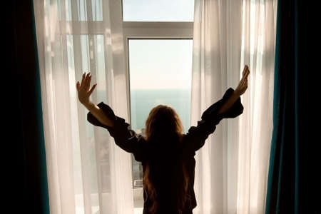 silhouette of a girl in a black men's shirt on the background of a window overlooking the ocean, early morning on vacation, meeting the dawnの写真素材