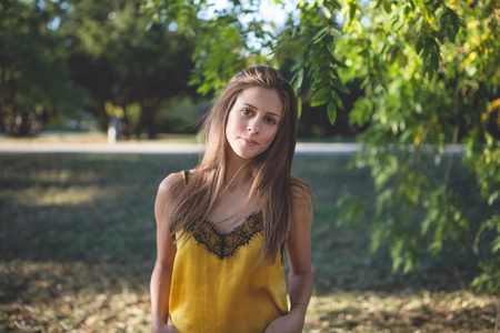 Beautiful girl with long healthy dark blonde hair in a yellow silk top in an autumn park, smiling at the cameraの写真素材