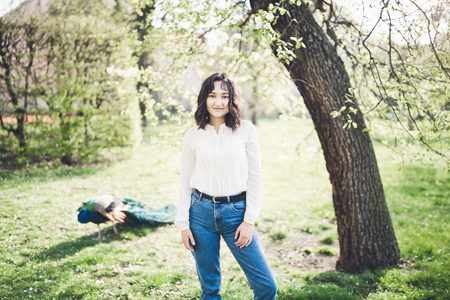 Happy asian young woman in a white blouse and jeans in the garden, peacock in the background. Springtime.の写真素材
