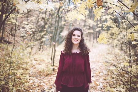 Beautiful girl with curly dark hair in a maroon top in an autumn park, smiling at the camera
