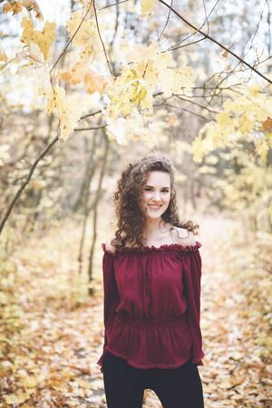 Beautiful girl with curly dark hair in a maroon top in an autumn park, smiling at the camera