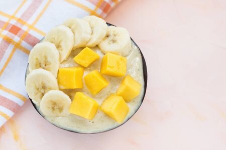 Yellow vegan homemade banana and mango dairy-free gelato ice cream in a coconut bowl with fresh pieces of mango and banana on top, pink background. Copy space, selective focus.の写真素材
