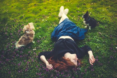 Beautiful redhead young woman lying on the grass, touching flowers, dogs at her sides. Outdoor...