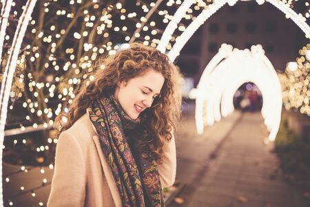 Beautiful curly hair smiling young woman in a coat outdoors with Christmas lights and decoration in...