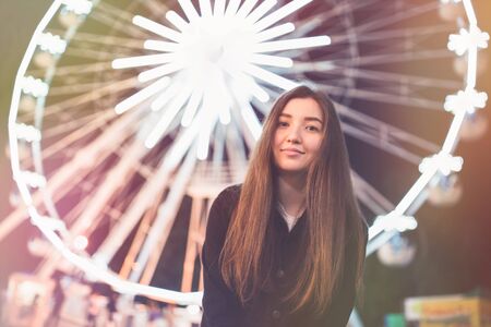 Beautiful girl in an amusement park at nght, smiling. Ferris wheel in the background. Copy space. Toned picture, soft focus.の写真素材