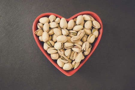 Roasted salted pistachios shelled in heart shaped red bowl on black background, concept of healthy eating vegan food and snack. Close up, selective focus, copy spaceの写真素材