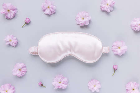 Pink satin eye sleeping mask and fresh spring Japanese cherry blossom flowers on pastel background. Insomnia, sleeplessness disorder, beauty sleep concept. Close up, copy space, flat lay. Accessories for girls and womenの写真素材