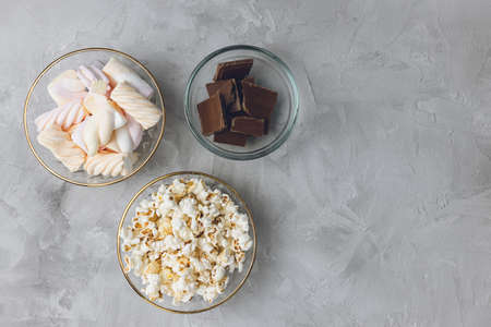 Popcorn, colorful pastel marshmallow, pieces of broken milk chocolate bar in glass bowls on gray...