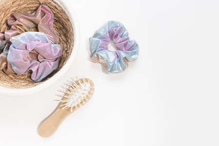 Trendy holographic iridiscent shiny metallic scrunchies in basket for accessories and small wooden bamboo hair brush on white background. Diy accessories, fashion and lifestyle concept. Top view, closeup, copy spaceの写真素材