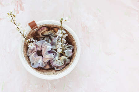 Trendy holographic shiny metallic scrunchies in basket for accessories and white cherry blossom flowers on pink background. Diy accessories, spring fashion and lifestyle concept. Top view, closeup, copy spaceの写真素材