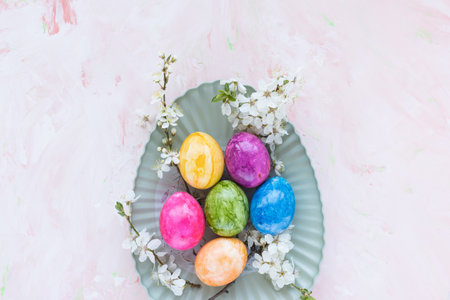 Colorful Easter eggs on plate and spring white cherry blossom flowers on pink background. Easter...