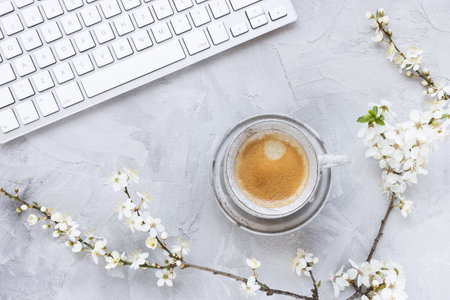 Computer keyboard on spring gray background with white cherry blossom flowers and morning espresso...