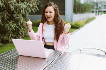 Young confident woman in casual clothes smile, raise her hands and celebrating work success while sitting and working on laptop in street cafe restaurant on summer day. Freelance, online business, online work, win, luck, self-employment. Copy spaceの写真素材
