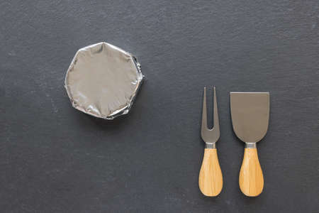 French round camembert cheese in wrapping foil paper and cheese knives on black slate plate....