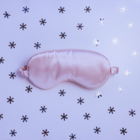 Pink satin eye sleeping mask on winter with glitter snowflakes background. Insomnia, good sleep and sleeplessness disorder concept. Accessories for girls and womenの写真素材