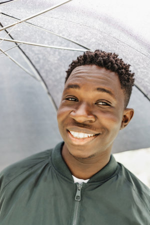 Young African American man with umbrella, under rain, smiling on street outdoor. Fall or spring...