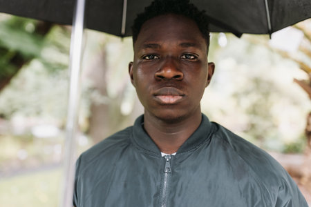 Young serious or sad African American man with umbrella, under rain on street outdoor. Fall or...