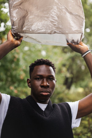 Young African American man outdoor with trash bag over his head, street portrait. Plastic pollution,...