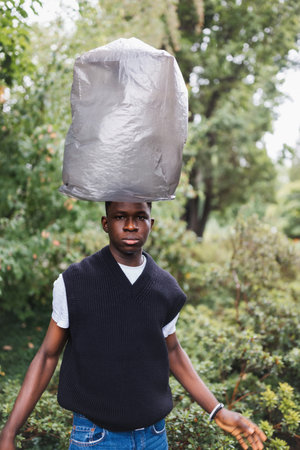Young African American man outdoor with trash bag over his head, street portrait. Plastic pollution, ecology, environmental conversation, mental health concept. Fall or spring seasonの写真素材