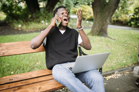 Excited Black Man Celebrating Success on Phone with Laptop in Park.の写真素材