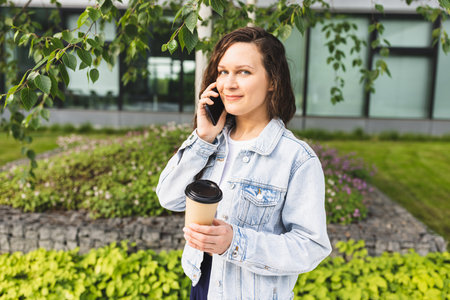 Woman Enjoying Coffee Outdoors Talking on Phone in Urban Green Space.