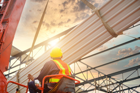 Construction workers are driving cranes to use cranes to lift large new roof sheets to install roofs on steel roof structures.の写真素材