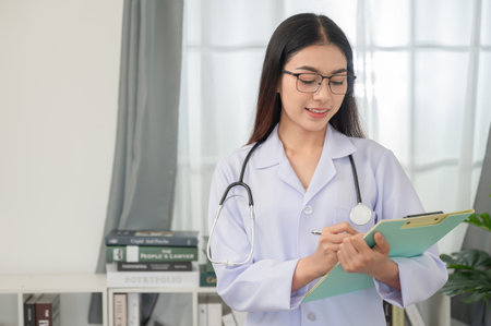 Portrait of a young Asian female doctor wearing glasses Standing with a clipboard in her office at the clinic.の写真素材