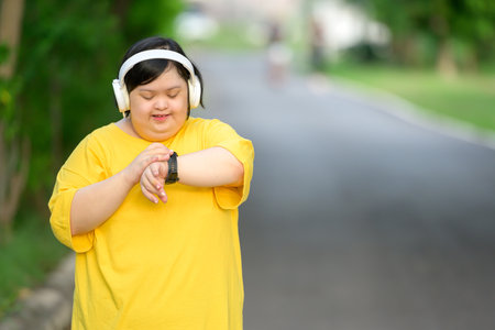 Asian girl with down syndrome wearing headphones and looking at smartwatch smiling happily walking to burn fat for exercise in a park in the middle of natureの写真素材