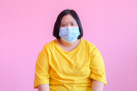 Image of a young woman with Down syndrome Wear a mask to protect against the COVID-19 coronavirus. She is a cerebral palsy student called Down syndrome. She wore a yellow dress on a pink background.の写真素材